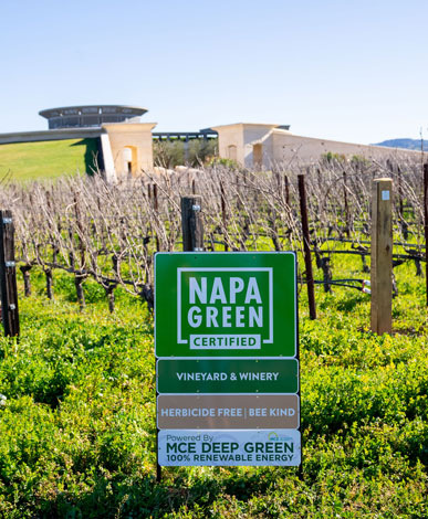 A Napa Green sign mentioning our sustainability certification, for the vineyard and winery, being herbicide and bee free, and using MCE deep green energy. The vines and winery are in the background.